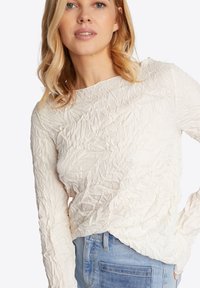 Gekreukte lange mouwen top in off-white stof met een ronde hals, gecombineerd met blauwe denim jeans, die een gestructureerd uiterlijk toont.