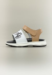 Παιδικό σανδάλι με λευκό μπροστινό λουράκι που φέρει μαύρο κείμενο και μπεζ λουράκι αστραγάλου με κλείσιμο Velcro σε λευκή και μαύρη σόλα.