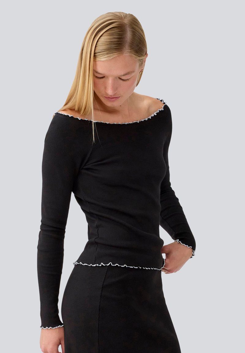 Schwarzes, geripptes Langarmoberteil mit einem schulterfreien Ausschnitt und weißem, Rüschensaum an den Ärmeln und dem Saum. Die schmale Passform betont die Silhouette.