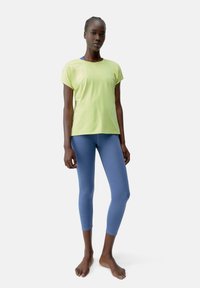 Lāmu zaļš īsām piedurknēm tops, kas kombinēts ar cieši pieguļošām, potīšu garuma gaiši ziliem legingiem, abi izgatavoti no gludas, vieglas auduma.