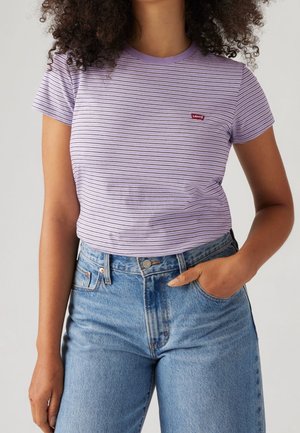Femme portant un t-shirt Levi's à rayures lilas avec col rond, rentré dans un jean bleu taille haute, une main dans la poche, cheveux bouclés visibles.