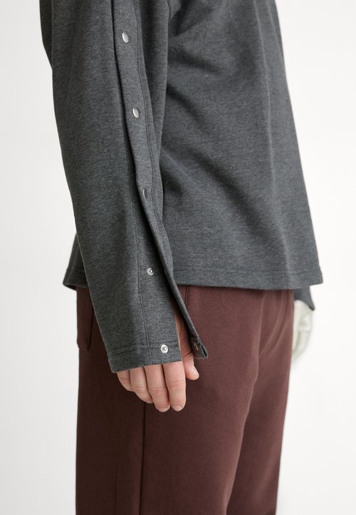 Pilkas ilgomis rankovėmis marškinis su apvaliu iškirpimu, turintis spaustukus ant rankovių ir lygų paviršių, derinamas su rudomis kelnėmis.