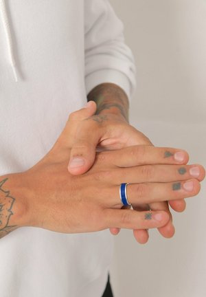 Blauwe en zilveren ring aan een hand met tatoeages, met een slank ontwerp en een gladde textuur. De handen zijn gepositioneerd met elkaar overlappende vingers.