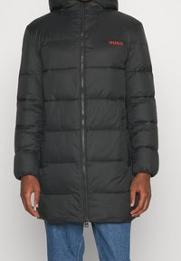 Schwarze Steppjacke mit Kapuze, verfügt über horizontale Steppnähte, einen Frontreißverschluss und ein rotes "HUGO"-Logo auf der Brust.