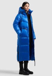 Ilgas mėlynas pūkinis paltas su gaubtu, blizgiu paviršiumi, siuvinėtu raštu ir užtrauktukais kišenėse, derinamas su juodais kulkšnies auliniais.