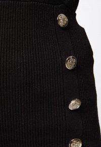 Close-up van zwarte ribbelstof met vier verticale, glanzende zilveren knopen met ingewikkelde wapenschildontwerpen.