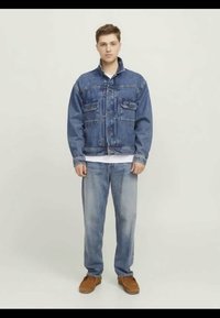 Casaco de ganga com dois bolsos no peito, jeans azul claro e calçado castanho. Apresenta um colarinho clássico e fecho com botões.
