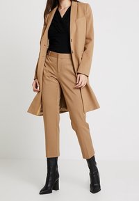 Kvinde iført en kamelfarvet frakke, matchende bukser, sort top og sorte hæle boots, der står mod en ensfarvet hvid baggrund.
