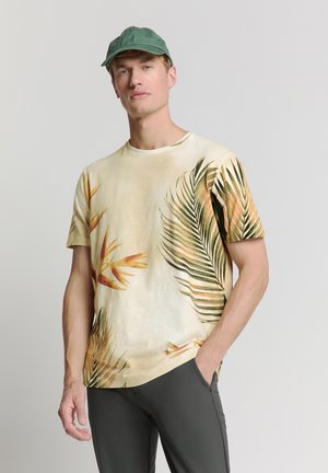 Kurzärmliges, helles T-Shirt mit tropischen Blattmustern in Grün und Orange. Kombiniert mit dunkelgrünen Hosen und einer passenden Kappe.