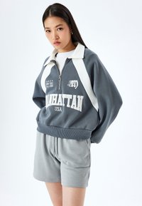 Felpa grigia corta con accenti bianchi, colletto a mezza zip e grafica frontale con scritto "MANHATTAN USA." Abbinata a pantaloni corti grigi chiari.