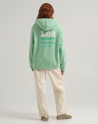 Persona con capelli rossi che indossa un hoodie mint green di Lee Crayola Magic Mint e pantaloni a gamba larga color crema, in piedi di fronte.