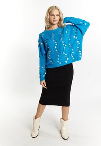Fényes kék, oversized pulóver texturált részletekkel és kör alakú flitterekkel, fekete kötött ceruzaszoknyával és krémszínű cowboy csizmával párosítva.