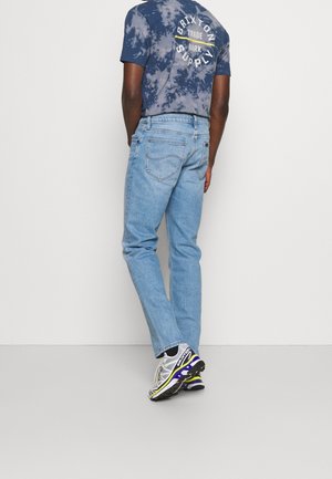 Man draagt lichtblauwe spijkerbroek, een blauw tie-dye Brixton Supply T-shirt en witte sportschoenen met paarse en gele accenten.