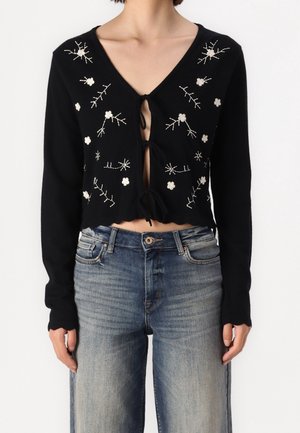 Maglione nero accorciato con maniche lunghe, scollo a V e ricamo floreale. Presenta una cintura frontale e orlo smerlato. Abbinato a jeans a vita alta.