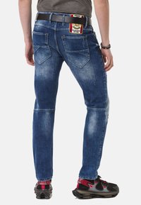 Blaue Jeans aus Denim mit einem ausgewaschenen Finish, fünf Taschen und einem Etikett auf der Rückseite. Getragen mit schwarzen Sneakers mit roten Akzenten.