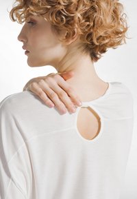 Osoba o kręconych, rudych włosach, ubrana w biały top z wycięciem na plecach, trzymająca lewy bark prawą ręką, widziana z boku i z tyłu.