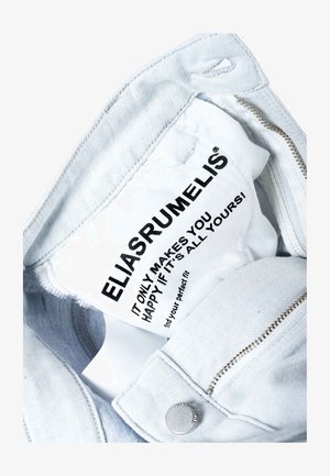 Primo piano di jeans in denim azzurro chiaro che mostrano un'etichetta interna bianca con il nome del marchio "ELIASRUMELIS" e una chiusura lampo.