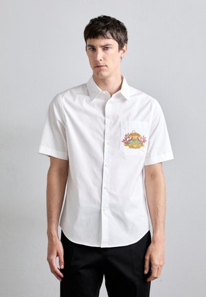 Chemise blanche à manches courtes en coton, dotée d'une poche sur la poitrine gauche avec un design brodé coloré, d'une fermeture à boutons sur le devant et d'un ourlet arrondi.