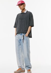 Schwarzes Kurzarmhemd mit strukturiertem Design, über einem gestreiften Hemd mit Kragen getragen. Hellblaue weite Jeans mit buntem Schlüsselanhänger-Detail. Rosa Mütze.