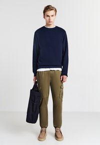 Felpa navy a maglia, indossata sopra una T-shirt bianca. Pantaloni cargo verde oliva con tasche laterali. Scarpe beige. Borsa a tracolla nera in tessuto.