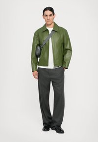Chaqueta de cuero verde sobre una camisa blanca, combinada con pantalones de pierna ancha grises y zapatos negros. Se lleva un pequeño bolso negro cruzado.