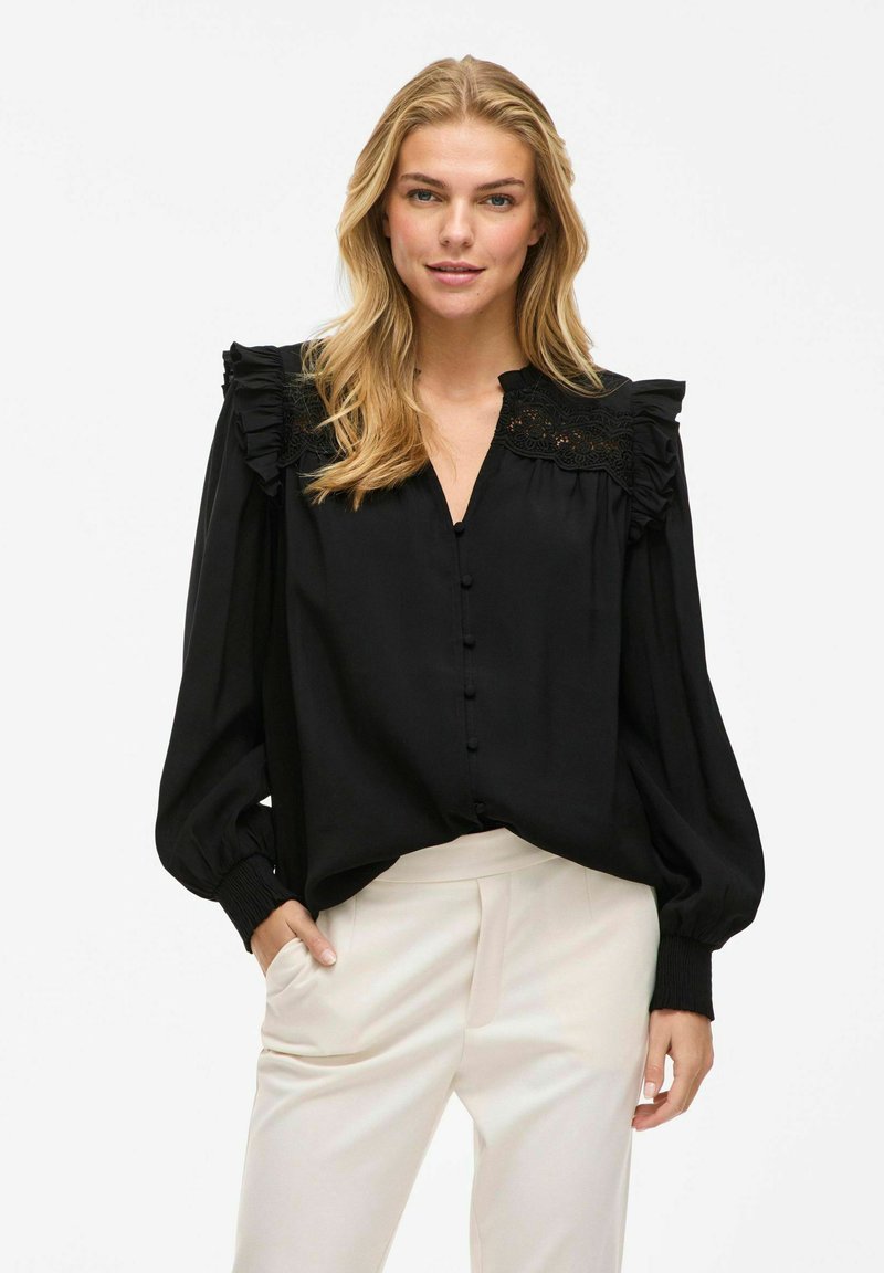 Blouse noire avec des accents en volants, fermeture à boutons à l'avant et empiècement brodé. Manches longues avec poignets ajustés. Le tissu semble lisse et léger.