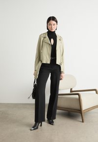 Jachetă de biker din piele verde deschis cu accente argintii, pulover negru cu gât înalt ce prezintă un decupaj, pantaloni negri cu talie înaltă și cizme cu vârful ascuțit până la gleznă.