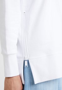 Sudadera blanca con cremallera frontal y puños acanalados, que destaca un zipper lateral y un dobladillo trasero ligeramente más largo, combinada con unos pantalones de denim azul claro.