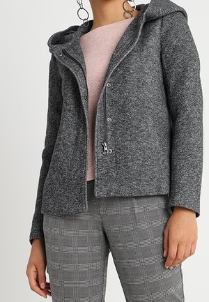 Vrouw die een grijze gestructureerde jas met ritssluiting en drukknopen draagt over een lichtroze top, gecombineerd met grijze geruite broek en grote hoepeloorbellen.