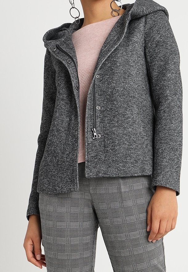 Femme portant une veste grise texturée zippée et à boutons-pression sur un haut rose clair, assortie à un pantalon gris à carreaux et de grandes boucles d'oreilles créoles.