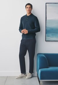 Dark teal knit polo shirt with three buttons, paired with dark trousers and light beige sneakers. Standing against a white wall.