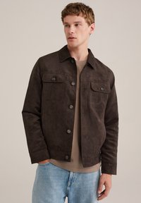 Chaqueta de ante marrón con un cuello clásico, que presenta dos bolsillos en el pecho y cierres de botón, combinada con una blusa beige y jeans azul claro.