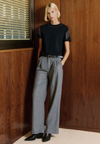 Camiseta negra con detalles de flecos en los hombros, pantalones amplios de tiro alto grises, botines negros con estampado, frente a paneles de madera.