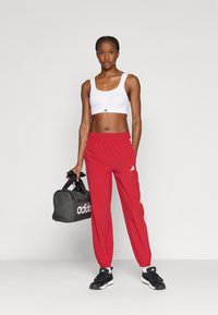 Κόκκινο αθλητικό παντελόνι με ελαστικές μανσέτες, λευκό αθλητικό σουτιέν και μαύρα αθλητικά παπούτσια. Κρατώντας μια γκρι τσάντα duffel με λευκές λογότυπες λεπτομέρειες.