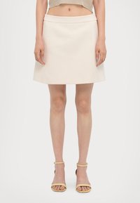 Une jupe A-line beige clair avec une texture lisse, dotée d'une large ceinture et de coutures latérales discrètes, assortie de sandales jaunes à lanières.