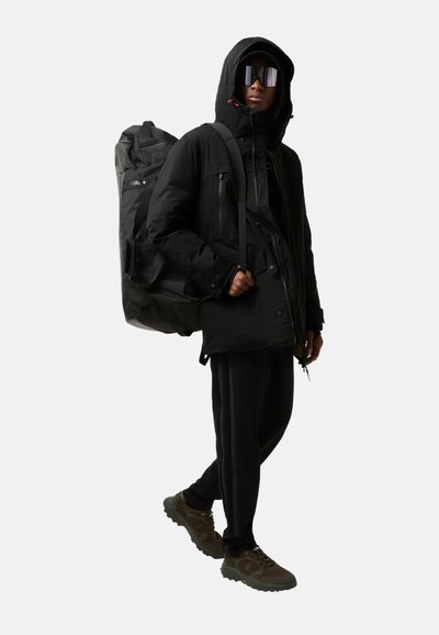 Černá péřová bunda s kapucí, se spoustou kapsářů. Kombinována s černými sportovními kalhotami a zelenými turistickými botami. Velký černý batoh.