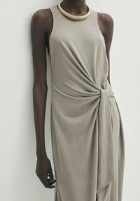 Robe sans manches, longueur genoux en tissu gris clair avec un détail de nœud torsadé à la taille. Accentuée par un collier en ton doré.