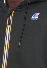 Svart zip-up hettegenser med en teksturert finish, med en livlig flerfarget stripe nedover midten og en K-Way-logo på brystet.
