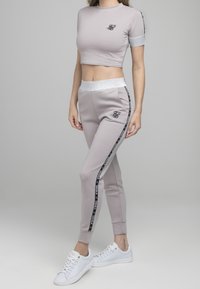 Camiseta corta de color gris claro con logotipo negro, acompañada de pantalones joggers a juego con franjas laterales negras y cinturilla reflectante plateada. Zapatillas blancas.