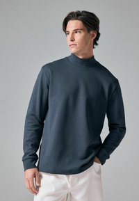 Mørkegrå langærmet top med høj krave, glat tekstur, afslappet pasform og synlige syninger, parret med lyse bukser.