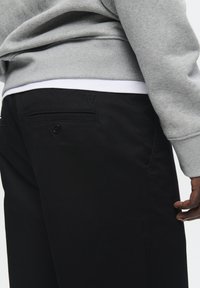 Black pants with a smooth texture, featuring a back pocket with a button and an embroidered "W" logo. Grey sweatshirt visible above.