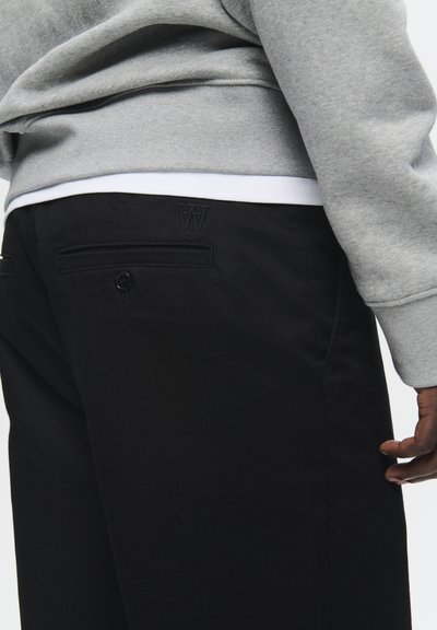 Sorte bukser med en glat tekstur, der har en baglomme med en knap og et broderet "W" logo. Grå sweatshirt synlig ovenfor.