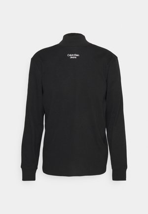 Zwart shirt met lange mouwen en opstaande kraag met het witte "Calvin Klein Jeans"-logo gecentreerd nabij de bovenrug.