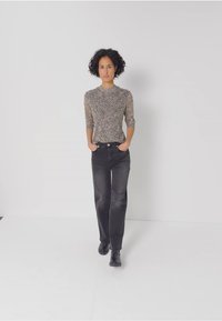 Musteroberteil mit langen Ärmeln in Beige mit dunklen Sprenkeln, kombiniert mit hochgeschnittenen schwarzen Jeans; beide figurbetont, Model steht mit Händen in den Taschen.