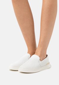 Scarpe bianche senza lacci con un motivo testurizzato, caratterizzate da una parte superiore liscia, punta rotonda e un piccolo accentuazione metallica sul lato.