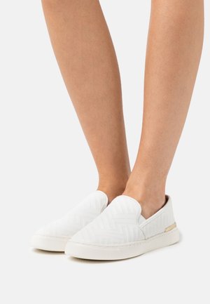 Baskets blanches à enfiler avec un motif texturé, présentant un dessus lisse, un bout arrondi et un petit accent métallique sur le côté.