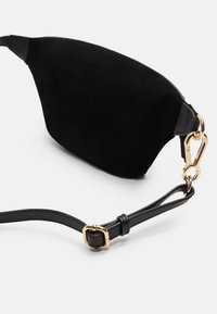 Bolso de piel de gamuza negra con forma curva, detalles de metal dorado y una correa de cuero negro con hebilla ajustable.