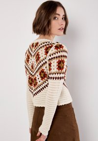 Gehäkelter Pullover mit einer Cremegrundlage, der ein geometrisches Muster in Orange, Braun und Grün aufweist. Lange Ärmel mit gerippten Bündchen.