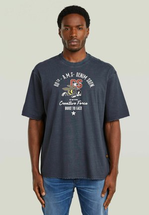 Man draagt een donkerblauw oversized T-shirt met bijenprint en tekst "GS Creative Force Built to Last," gecombineerd met een blauwe spijkerbroek, staand voor een neutrale achtergrond.