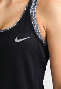 Zwarte sportieve tanktop met een zilveren Nike-logo, voorzien van een ronde halslijn en dunne bandjes. Gelaagd met een gemêleerd zwart-witte top eronder.